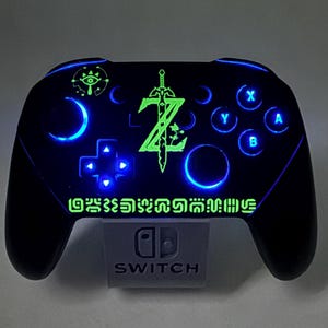 May include: A black wireless game controller featuring glowing blue and green light accents. The center displays a green Zelda logo with a sword, a Sheikah eye symbol, and glowing blue buttons. A row of green Hylian text is printed below the buttons.
