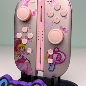 May include: Pink and silver Nintendo Switch Joy-Con controllers featuring Princess Peach artwork. The controllers have pink buttons and accents, with a star and bow pattern. The controllers are displayed on a purple and blue stand with the text "Game Cosmos".