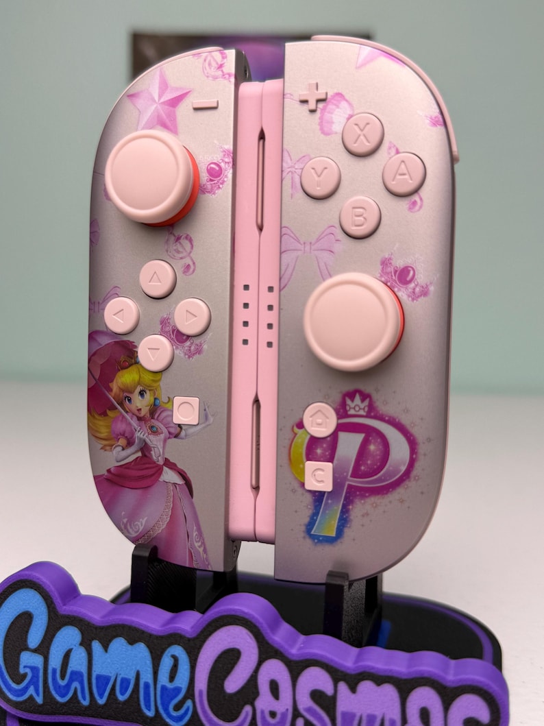 May include: Pink and silver Nintendo Switch Joy-Con controllers featuring Princess Peach artwork. The controllers have pink buttons and accents, with a star and bow pattern. The controllers are displayed on a purple and blue stand with the text "Game Cosmos".