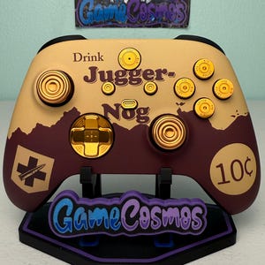 以下が含まれることがあります： タンとマルーンのデザインのカスタムビデオゲームコントローラー。「Drink Juggernog」の文字と弾丸型のボタンが付いています。スタンドには、青と紫で「Game Cosmos」の文字が書かれています。