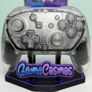 Könnte beinhalten: Ein transparenter kabelloser Videospiel-Controller mit einer klaren Kunststoffhülle, zwei Analogsticks, einem Steuerkreuz und vier Aktionstasten. Der Controller steht auf einem schwarzen Ständer mit einem violett-blauen GameCosmos-Logo.