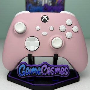 May include: A pastel pink wireless gaming controller featuring white analog sticks, a white directional pad, and white action buttons labeled A, B, X, and Y. The controller rests on a black display stand with a purple and blue GameCosmos logo.