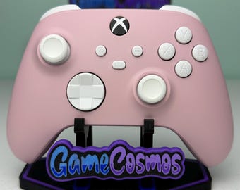 May include: A pastel pink wireless gaming controller featuring white analogue sticks, a white directional pad, and white action buttons labelled A, B, X, and Y. The controller rests on a black display stand with a purple and blue GameCosmos logo.