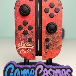 May include: A pair of video game controllers featuring a red, textured, rusted-metal pattern with the text Nuka Cola printed in yellow on the left side. The controllers are mounted on a black stand with a purple and blue GameCosmos logo.