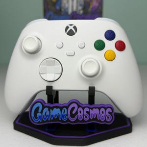 May include: A white wireless video game controller with two white joysticks, a white directional pad, and four colored action buttons in green, blue, yellow, and red. It rests on a black stand featuring a purple and blue GameCosmos logo.