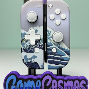 May include: A pair of Nintendo Switch Joy-Con controllers with a wave design. The controllers are mounted on a stand with the text "Game Cosmos" in blue and purple. The controllers have white buttons and a light purple background.