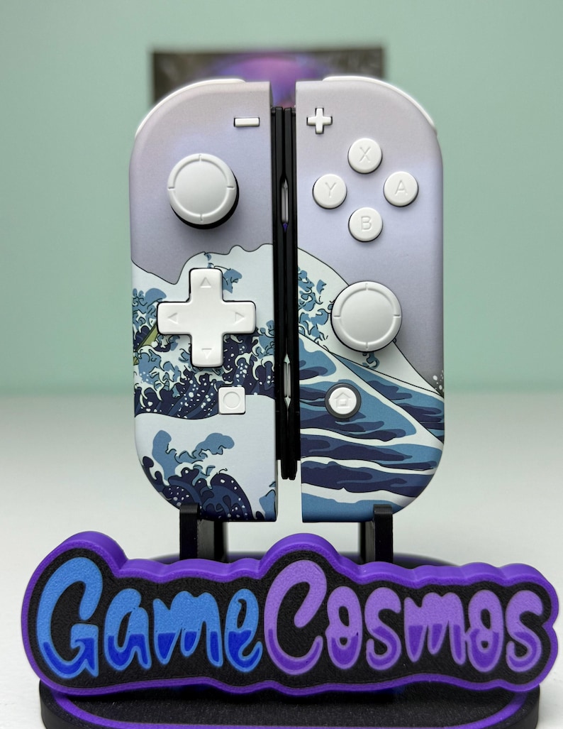 May include: A pair of Nintendo Switch Joy-Con controllers with a wave design. The controllers are mounted on a stand with the text "Game Cosmos" in blue and purple. The controllers have white buttons and a light purple background.