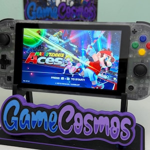 May include: A clear, handheld gaming console displaying "Mario Tennis Aces" on its screen, resting on a black stand with a purple and blue "Game Cosmos" logo. The console has colorful buttons and a black D-pad.