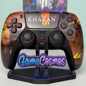 May include: A black and brown video game controller featuring the text "THE FIRST BERSERKER KHAZAN" and a character graphic. The controller rests on a stand with the text "Game Cosmos".
