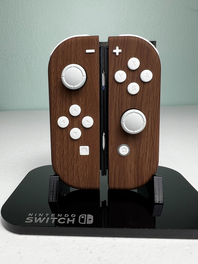 May include: A brown and white Nintendo Switch OLED console with a wood grain design on the controllers. The console is on a black stand with the Nintendo Switch logo.