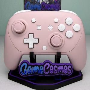 Puede incluir: Un mando de juego inalámbrico rosa con botones blancos y una cruceta blanca. El mando está sobre un soporte negro con el texto "Game Cosmos" en azul y morado. El soporte tiene una base negra.