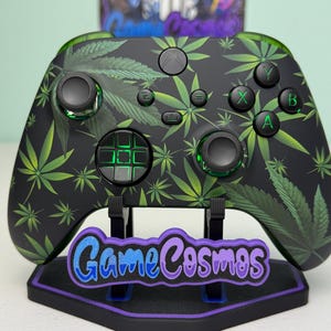 May include: Black Xbox controller with a green cannabis leaf pattern. The controller has green accents on the buttons and joysticks. It is displayed on a stand with the text "Game Cosmos" in blue and purple.