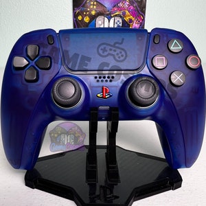 Puede incluir: Un mando de juego PlayStation 5 DualSense azul con un acabado translúcido se muestra en un soporte negro. El mando tiene un logotipo de PlayStation negro en la parte delantera y botones negros.