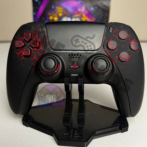 Pode incluir: Um comando PlayStation 5 preto com detalhes vermelhos e um padrão de favo de mel nas costas. O comando está em um suporte preto com o logotipo do PlayStation.