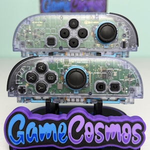 Puede incluir: Dos mandos de videojuego de plástico transparente con circuitos impresos verdes visibles, botones negros y joysticks negros. Los mandos están apilados verticalmente y cuentan con crucetas, botones de acción y botones de sistema.