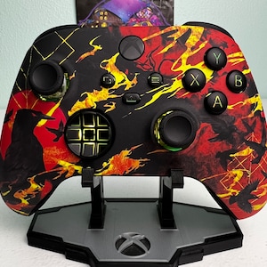 May include: A black and red Xbox controller with a fiery design. The controller has a black and white button pad and a black and white D-pad. The controller is on a black stand.