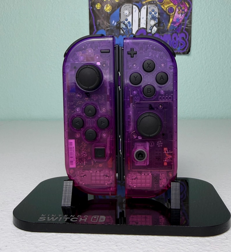 May include: A pair of transparent purple Nintendo Switch Joy-Con controllers with black buttons and joysticks. The controllers are mounted on a black stand with the words "NINTENDO SWITCH" printed on the front.