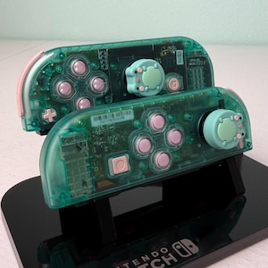 May include: Two teal green Nintendo Switch controllers with pink buttons and light green and pink animal-shaped joystick caps. The controllers are on a black stand with the words "Nintendo Switch" printed on the front.