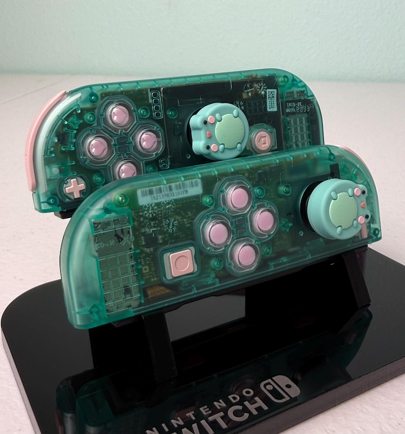 May include: Two teal green Nintendo Switch controllers with pink buttons and light green and pink animal-shaped joystick caps. The controllers are on a black stand with the words "Nintendo Switch" printed on the front.