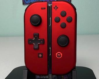Może przedstawiać: Para metalicznych czerwonych kontrolerów Joy-Con do konsoli Nintendo Switch zamocowanych na czarnej stacji ładującej. Lewy kontroler posiada krzyżak, joystick i przycisk minus. Prawy kontroler ma joystick, cztery przyciski akcji, przycisk plus oraz przycisk home z podświetlanym na czerwono pierścieniem. Napis GameCosmos jest widoczny na znaku w tle.