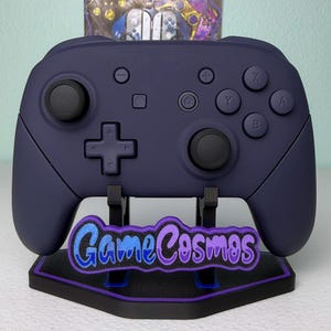 May include: A dark purple wireless video game controller rests on a black display stand. The stand features a purple-outlined logo plate with the text GameCosmos in blue and purple lettering. The controller has black joysticks.