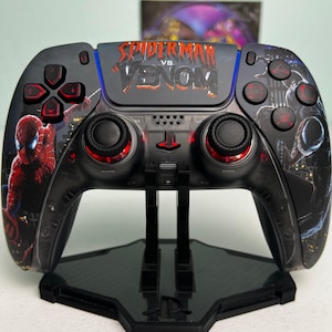 May include: A black wireless game controller featuring a Spider-Man vs Venom graphic on the center touchpad. The left side displays Spider-Man, while the right side shows Venom. The controller includes red accents on the buttons and joysticks.