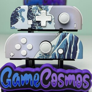 May include: Two gray and white game controllers with a blue wave design, displayed on a black stand with the text "Game Cosmos" in blue and purple. The controllers have white buttons and a white directional pad.