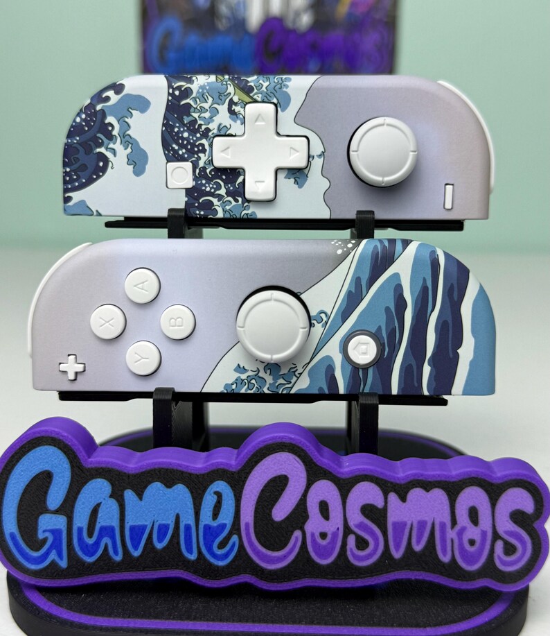 May include: Two gray and white game controllers with a blue wave design, displayed on a black stand with the text "Game Cosmos" in blue and purple. The controllers have white buttons and a white directional pad.