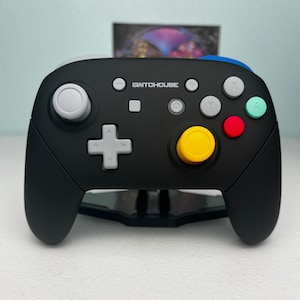 May include: A black wireless game controller featuring the text SWITCHCUBE in white. It includes a light gray D-pad, a white left joystick, a yellow right joystick, and colored action buttons in green, red, and gray on the right side.