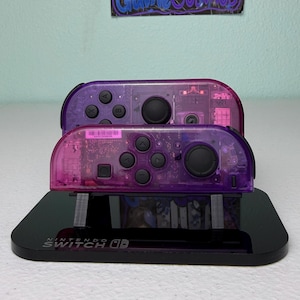 May include: Two transparent purple Nintendo Switch Joy-Con controllers stacked on a black stand. The controllers have black buttons and a clear casing, revealing internal components. The stand has the Nintendo Switch logo.