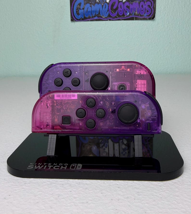 May include: Two transparent purple Nintendo Switch Joy-Con controllers stacked on a black stand. The controllers have black buttons and a clear casing, revealing internal components. The stand has the Nintendo Switch logo.