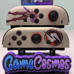 May include: Two gray game controllers with black buttons and red accents are stacked on a purple and blue stand. The top controller features a figure graphic, while the bottom one has a circular design. The stand reads "Game Cosmos."