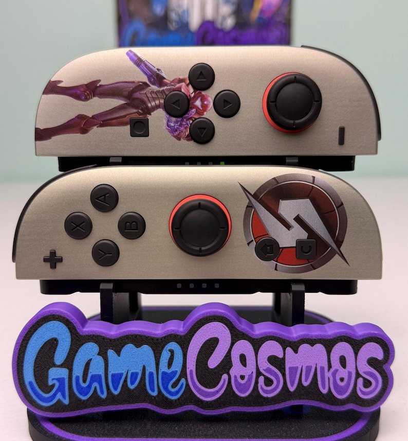 May include: Two gray game controllers with black buttons and red accents are stacked on a purple and blue stand. The top controller features a figure graphic, while the bottom one has a circular design. The stand reads "Game Cosmos."