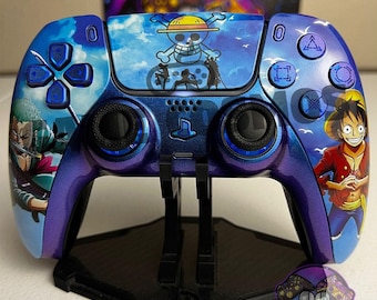 以下が含まれることがあります： 青と紫のワイヤレスビデオゲームコントローラー。ワンピースのアニメグラフィックが施されており、タッチパッドには麦わら帽子のドクロロゴ、サイドグリップにはルフィとゾロのキャラクターイラストが描かれ、青いボタンが付いています。