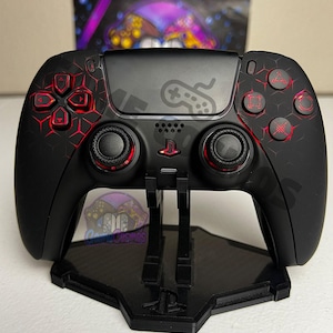 May include: A black wireless video game controller featuring a red geometric honeycomb pattern on the faceplate, red accents around the analog sticks, and a central PlayStation logo. The controller rests on a black plastic display stand.