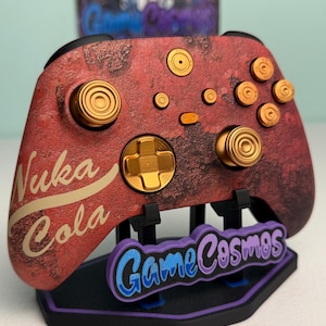 May include: A video game controller featuring a weathered red and brown textured finish with the text Nuka Cola printed in cream. The controller includes metallic gold buttons, joysticks, and a directional pad, resting on a black display stand.