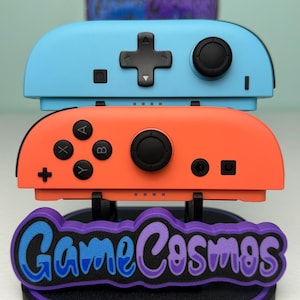 Può includere: Due controller di gioco, uno blu e uno arancione, appoggiati su un supporto con la scritta "Game Cosmos". I controller hanno pulsanti neri e pulsanti direzionali. Il supporto è nero con un bordo viola. Il testo è viola e blu.