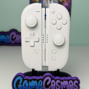 May include: A pair of white video game controllers featuring a directional pad, two analog sticks, four action buttons labeled X, Y, A, and B, plus home and capture buttons. The controllers are mounted on a black stand with a purple GameCosmos logo.