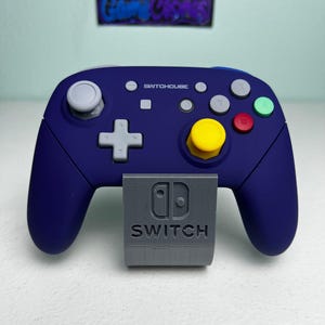 Könnte beinhalten: Ein dunkelvioletter kabelloser Gamecontroller mit dem in Weiß aufgedruckten Text SWITCHCUBE. Er verfügt über einen gelben Joystick, einen roten B-Knopf, einen grünen A-Knopf, graue Tasten und ein graues Steuerkreuz auf einem grauen Ständer mit Switch-Logo.