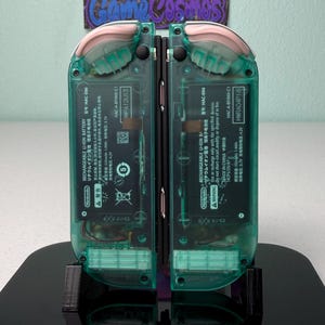 May include: Two teal green Nintendo Switch handheld gaming consoles with pink accents on the top and bottom of the device. The consoles are on a black stand with the Nintendo Switch logo.