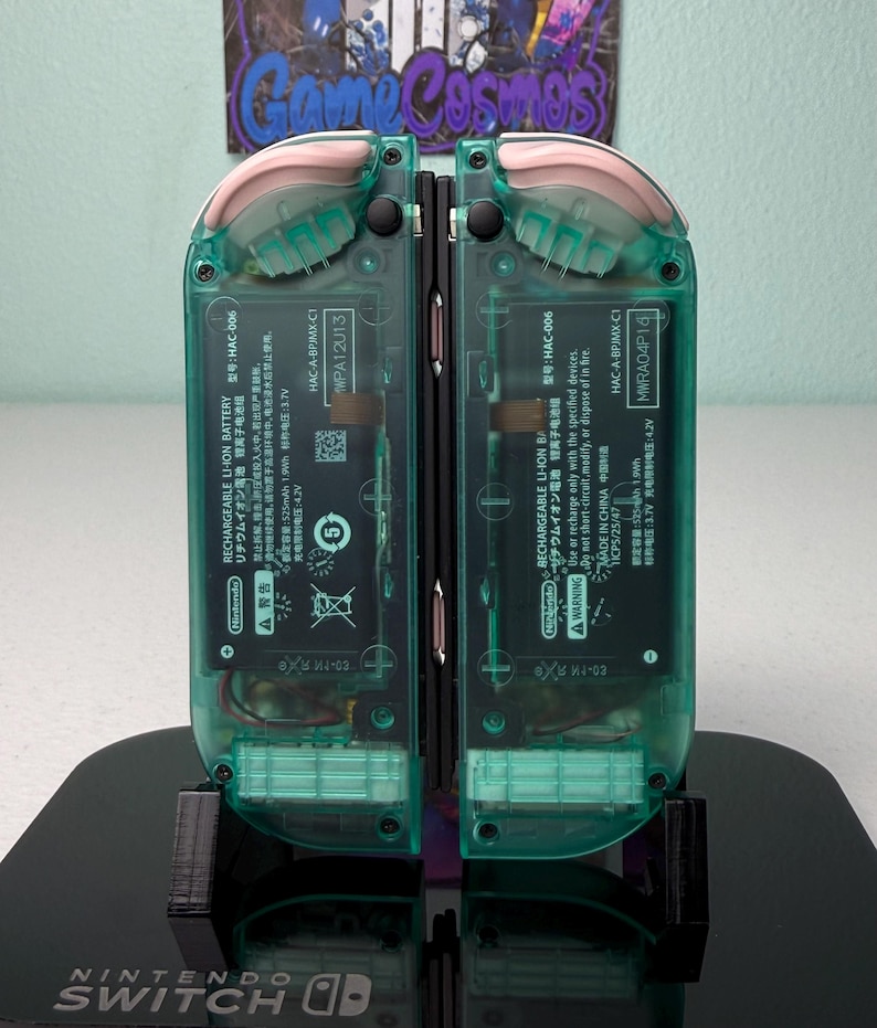 May include: Two teal green Nintendo Switch handheld gaming consoles with pink accents on the top and bottom of the device. The consoles are on a black stand with the Nintendo Switch logo.