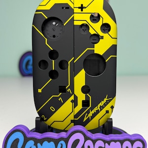 Puede incluir: Mando de juego negro y amarillo con tem&aacute;tica Cyberpunk 2077. El mando tiene un dise&ntilde;o de placa de circuito con el texto "2077" y "Cyberpunk 2077". El mando est&aacute; sobre un soporte con el texto "Game Cosmos".