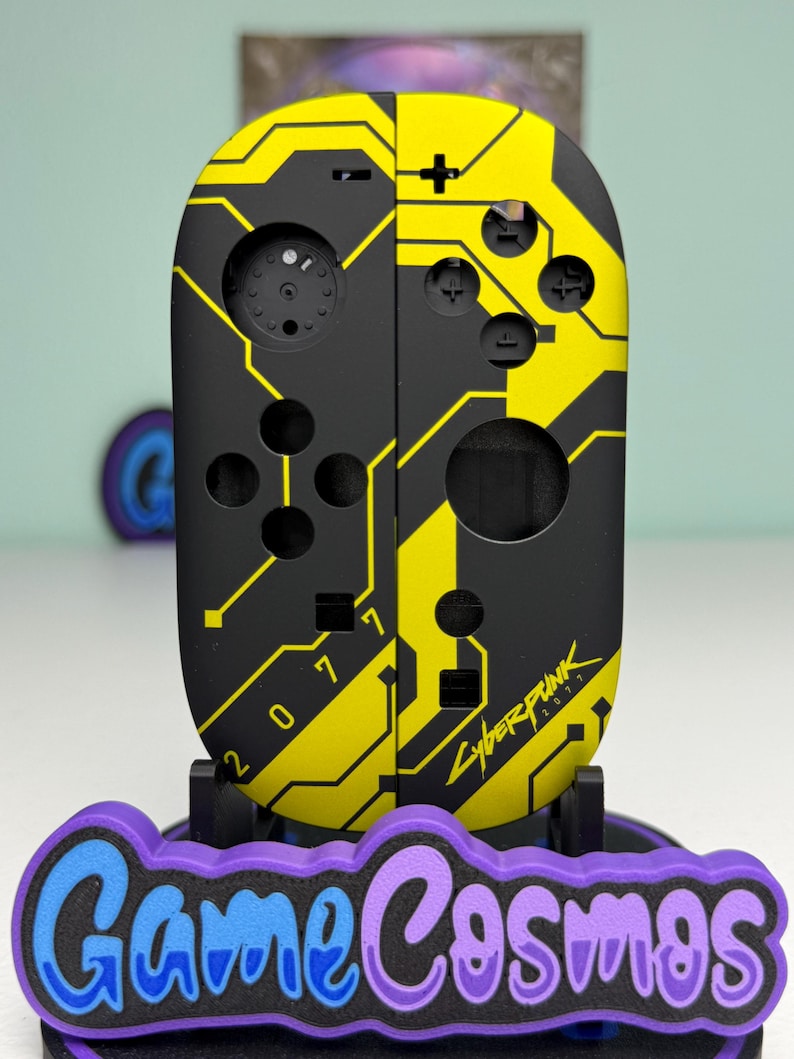 Puede incluir: Mando de juego negro y amarillo con tem&aacute;tica Cyberpunk 2077. El mando tiene un dise&ntilde;o de placa de circuito con el texto "2077" y "Cyberpunk 2077". El mando est&aacute; sobre un soporte con el texto "Game Cosmos".