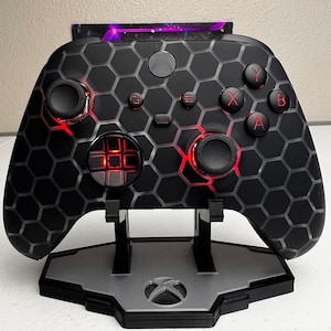 May include: A black Xbox controller with a honeycomb pattern and red buttons is displayed on a gray stand. The controller has a black and gray faceplate with a honeycomb pattern.