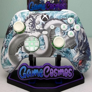 May include: Custom Xbox controller featuring a detailed dragon design in shades of gray, white, and blue. The controller has white joysticks and green-accented buttons. The controller is displayed on a black stand with the text "Game Cosmos" in purple and blue.