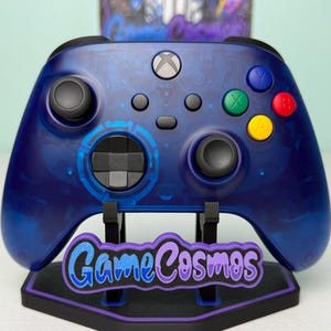 Puede incluir: Un mando de Xbox azul con un soporte a juego. El mando tiene botones de colores y un diseño transparente. El soporte presenta el texto "Game Cosmos" en una fuente colorida y estilizada. El tema general es el juego.