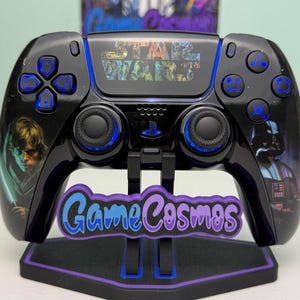 Puede incluir: Mando de videojuegos negro con temática de Star Wars, con detalles azules y gráficos de personajes. El mando se muestra en un soporte con el texto "Game Cosmos". El logotipo de Star Wars es visible.