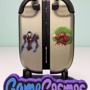 May include: A silver and black gaming console accessory with two stickers, one of a character in a suit and the other of a green and red creature. The accessory is on a purple and black stand with the text "Game Cosmos".