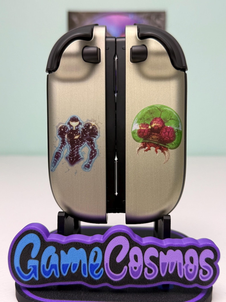 May include: A silver and black gaming console accessory with two stickers, one of a character in a suit and the other of a green and red creature. The accessory is on a purple and black stand with the text "Game Cosmos".