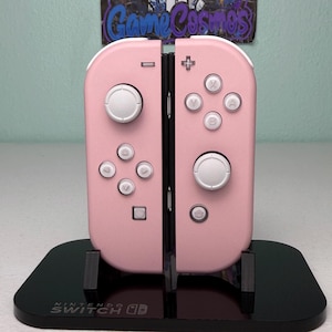 May include: A pair of pastel pink Nintendo Switch Joy-Con controllers with white buttons and joysticks, mounted on a black display stand. The base of the stand features the white Nintendo Switch logo. The controllers have standard directional inputs.